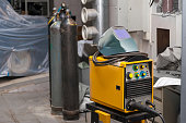 A yellow welding machine with a protective mask on top stands in a metal workshop near carbon dioxide cylinders. Industry and production. Construction works.