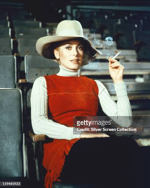 Catherine Deneuve, French actress, smoking a cigarette while wearing a wide-brimmed hat, a red sleeveless top and a white polo neck jumper, in a...