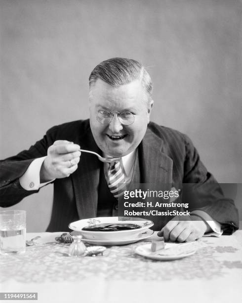 1930s MIDDLE-AGED MAN WEARING PINCE-NEZ EYEGLASSES PINSTRIPED SUIT LOOKING AT CAMERA SITTING AT DINING TABLE EATING BOWL OF SOUP