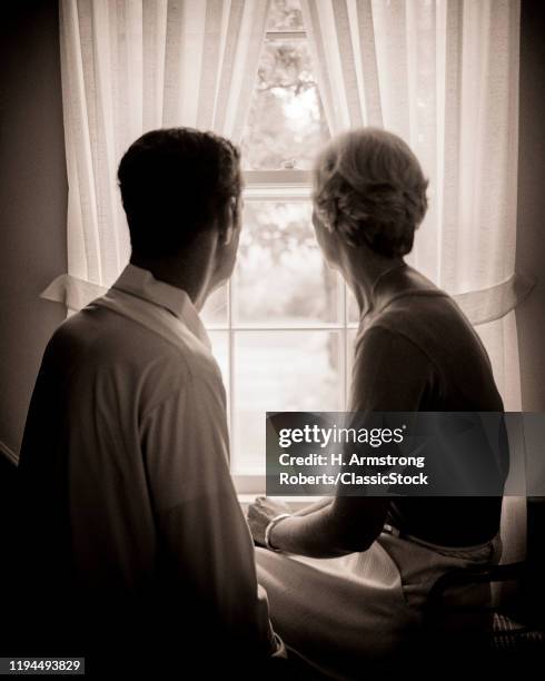 1970s ANONYMOUS SILHOUETTE OF MATURE MAN AND WOMAN LOOKING OUT OF WINDOW WITH TIE BACK CURTAINS