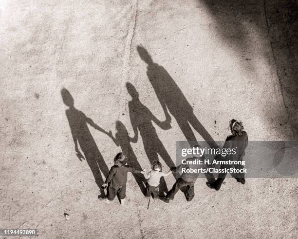 1950s OVERHEAD OF ANONYMOUS FAMILY OF FOUR STANDING HOLDING HANDS CASTING SHADOWS ON GROUND IN FRONT OF THEM