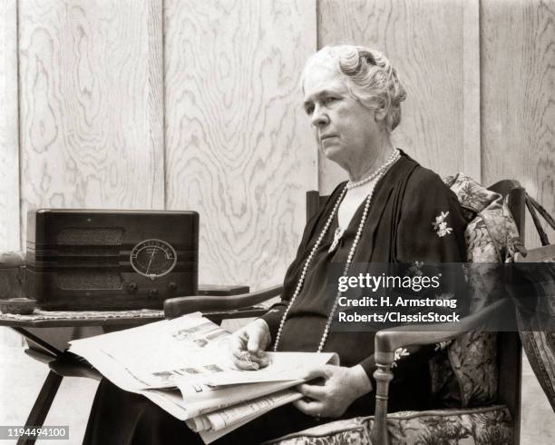 1930s 1940s SERIOUS WORRIED SENIOR WOMAN HOLDING NEWSPAPER IN HER LAP LISTENING TO RADIO