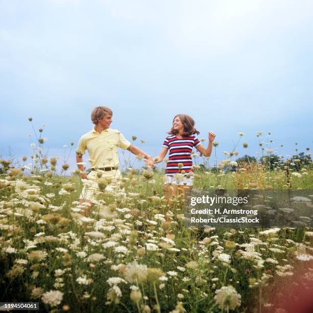 1970s TEENAGE COUPLE HOLDING HANDS RUNNING THROUGH A FIELD OF QUEEN ANNE'S LACE SMILING WEARING SUMMER CLOTHES