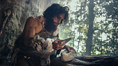 Primeval Caveman Wearing Animal Skin Holds Sharp Stone and Makes First Primitive Tool for Hunting Animal Prey, or to Handle Hides. Neanderthal Using Handax. Dawn of Human Civilization