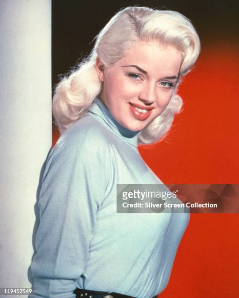 Diana Dors . British actress, wearing a light blue polo neck jumper in a studio portrait, against a red background, circa 1955.