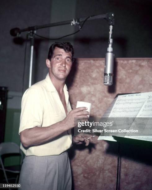 Dean Martin , US actor and singer, holding a cup and smoking a cigarette while standing before a sheet music stand and a microiphone in a recording...