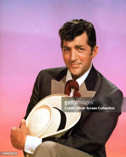 Dean Martin , US actor and singer, holding a cowboy hat and posing in costume for a studio portrait, against a pink background, 1960.