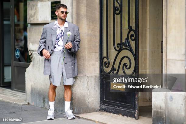 Jean-Sebastien Rocques wears sunglasses, a white shirt with navy blue prints, an oversized grey Prince of Wales long jacket, light grey shorts with...