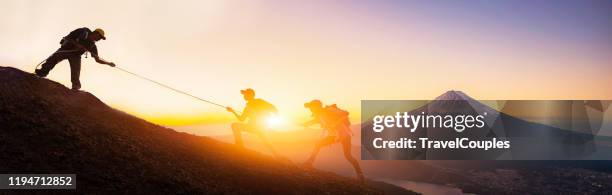group of travellers with backpacks over sunrise background. young asian three hikers climbing up on the peak of mountain near mountain fuji. people helping each other hike up. giving a helping hand. helps and team work concept - rope high rescue photos et images de collection
