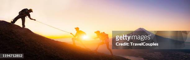 group of travellers with backpacks over sunrise background. young asian three hikers climbing up on the peak of mountain near mountain fuji. people helping each other hike up. giving a helping hand. helps and team work concept - to pull together stock pictures, royalty-free photos & images