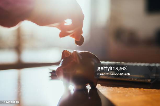 hand and a piggy bank and coin on a table in backlight. - sparen stock-fotos und bilder