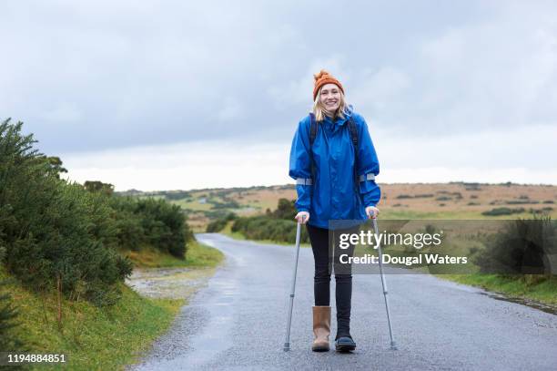 positive female hiker with crutches on a rural country lane. - crutch stock-fotos und bilder