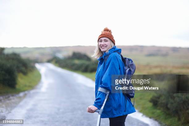 happy hiker with crutches walking in rain on rural country road. - crutches stockfoto's en -beelden