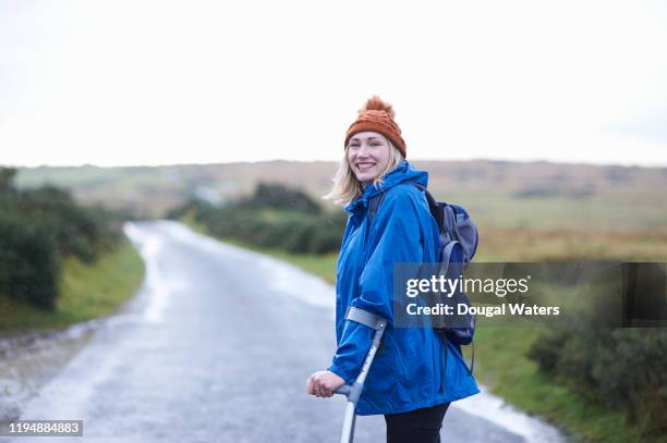 happy hiker with crutches walking in rain on rural country road. - crutch stock pictures, royalty-free photos & images