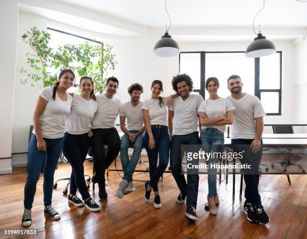 happy group of volunteers at an office all wearing white t-shirts smiling at camera - t shirt stock pictures, royalty-free photos & images