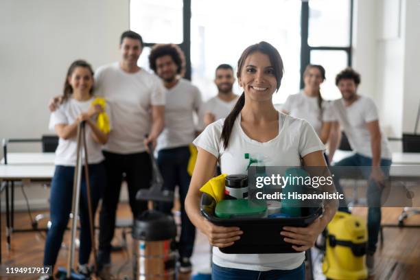 mooie schoonmakende vrouw met haar team bij een bureau dat een emmer met het schoonmaken van producten houdt die allen bij camera glimlachen - office cleaning stockfoto's en -beelden