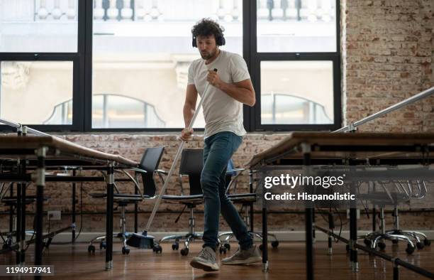 young male janitor cleaning an office while listening to music with wireless headphones - cleaner man uniform stock pictures, royalty-free photos & images