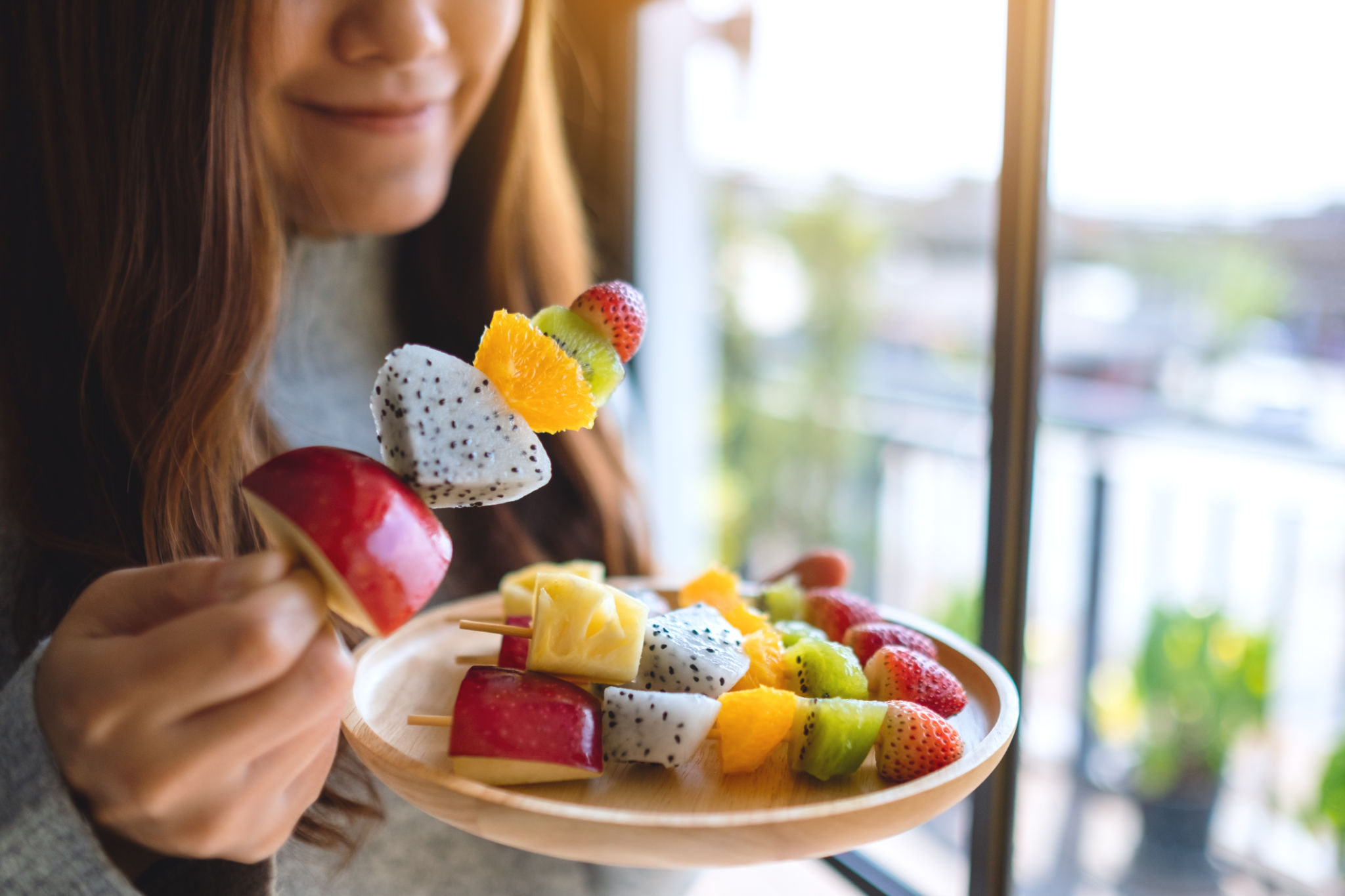 an asian woman holding and eating a fresh mixed fruits on skewers an asian woman holding and eating a fresh mixed fruits on skewers