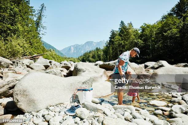 Father and daughter putting feet in river while having picnic during road trip