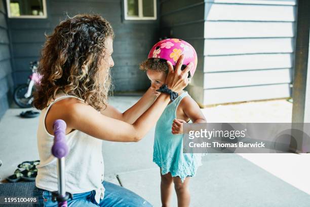 smiling mother helping toddler daughter put on helmet before riding scooter - protezione foto e immagini stock