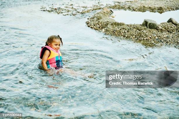 Smiling young girl wearing life jacket sitting in river on summer afternoon