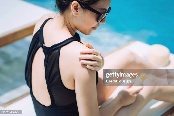 young asian woman applying suntan lotion while sunbathing by the swimming pool - asian woman swimsuit stock-fotos und bilder