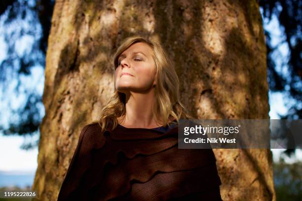 portrait serene woman with eyes closed standing at sunny tree trunk - forest bathing stock pictures, royalty-free photos & images