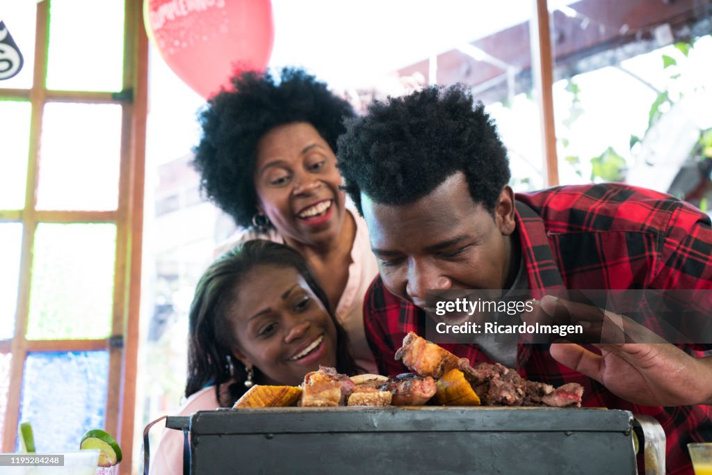 Latin man with afro hair and brown skin, approximately 29 years old, has a barbecue with meat of chorizo ​​sausage in front of him, looks at the camera and opens his big mouth while we look back at his wife and mother-in-law watching him