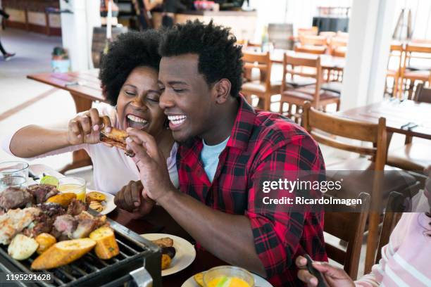 mujer latina con piel oscura y pelo afro de aproximadamente 50 años vestida casualmente está sentada en una mesa de restaurante donde almorza con su familia comida típica colombiana mientras su yerno hombre latino con piel morena y pelo afro la mira - 50 54 years fotografías e imágenes de stock