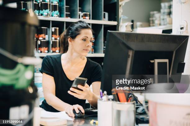 Sales woman using computer while holding mobile phone in store
