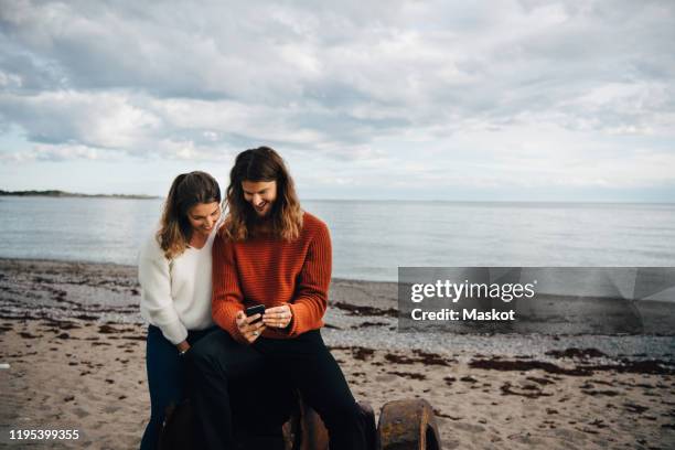 smiling man showing mobile phone to woman while sitting at beach - stockholm beach stock pictures, royalty-free photos & images