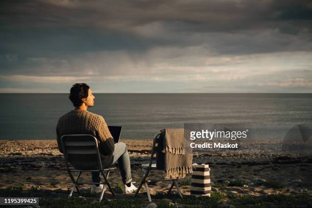 thoughtful man using laptop while sitting at beach against cloudy sky - beach seat stock pictures, royalty-free photos & images