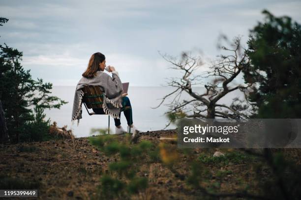 rear view of woman drinking coffee while sitting with laptop on chair against lake - mobile worker stock-fotos und bilder