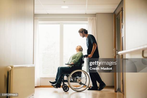 full length side view of male nurse pushing disabled senior woman on wheelchair in alley at retirement home - maatschappelijke dienstverlening stockfoto's en -beelden