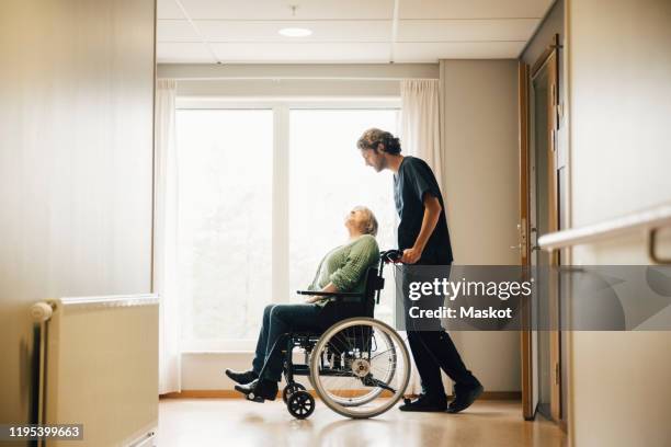 full length side view of male nurse pushing disabled senior woman on wheelchair in alley at retirement home - social worker stock pictures, royalty-free photos & images