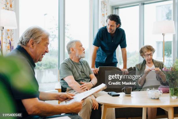 male healthcare worker talking to senior man sitting on sofa amidst friends at retirement home - retirement community stock pictures, royalty-free photos & images