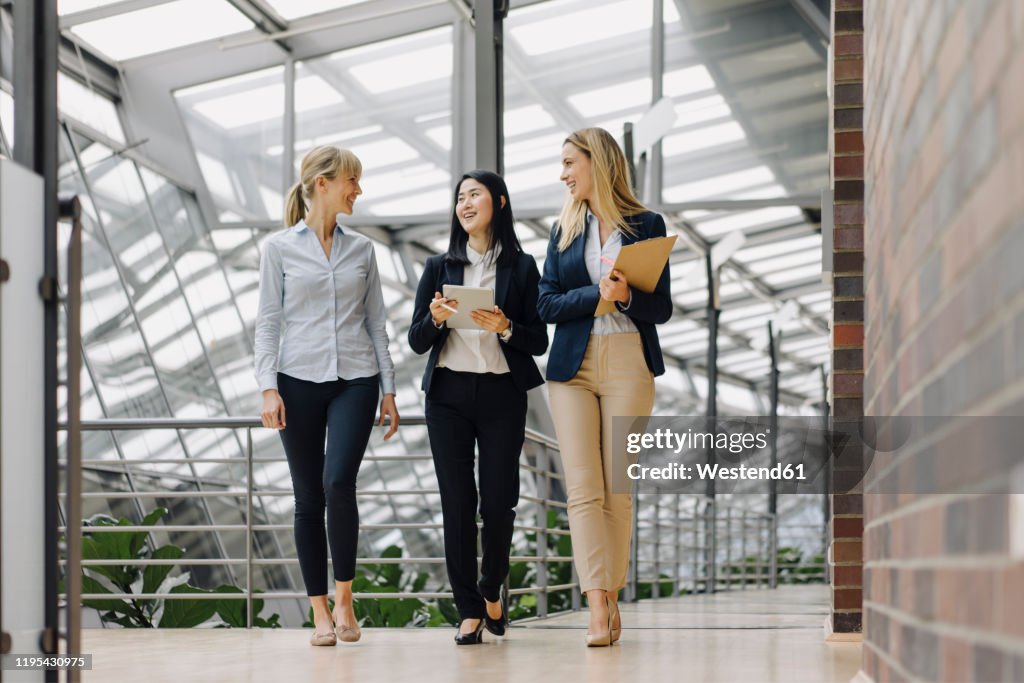 Three businesswomen with tablet walking and talking in modern office building