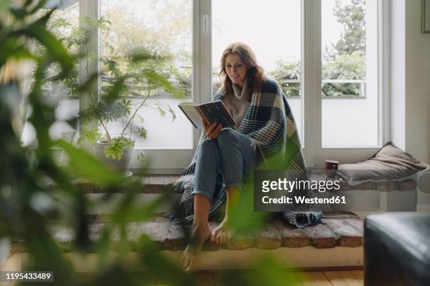 mature woman sitting on wondow sill, wrapped in blanket, reading book - escapism reading stock pictures, royalty-free photos & images