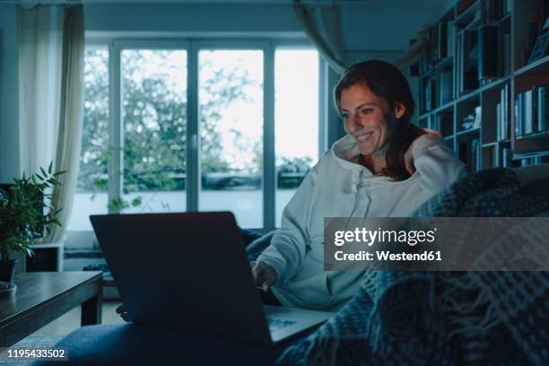 woman sitting on couch of dark living room, using laptop - living room dark stock-fotos und bilder