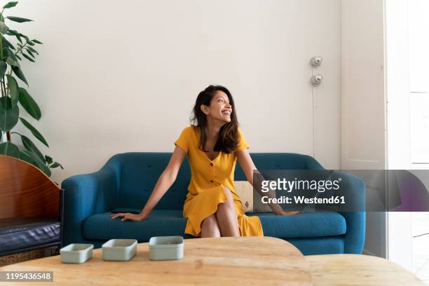 happy young woman sitting on couch looking sideways - yellow dress stock pictures, royalty-free photos & images