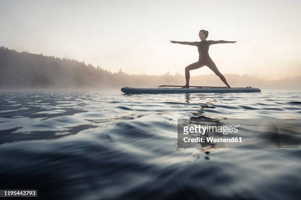 woman practicing paddle board yoga on lake kirchsee in the morning, bad toelz, bavaria, germany - paddleboard stock-fotos und bilder