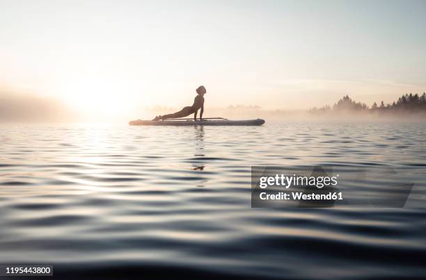 woman practicing paddle board yoga on lake kirchsee in the morning, bad toelz, bavaria, germany - sunrise yoga stock-fotos und bilder