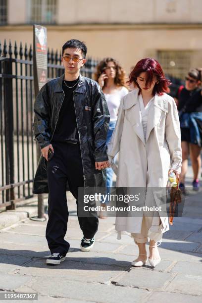 Guest wears sunglasses, a necklace, a black top, a shiny black trench coat, black full-length pants with thin white stripes, black sneakers ; A guest...