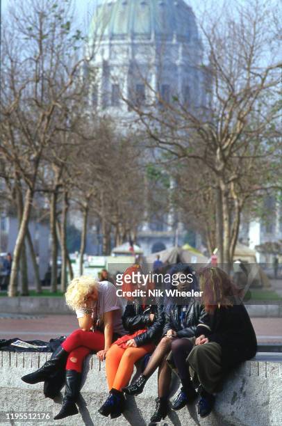 Girls in punk style colored hair sit smoking and chatting on wall in city square in front of the Capitol Building in downtown San Francisco circa 1980