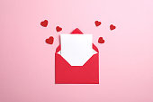 Red paper envelope with blank white note mockup inside and Valentines hearts on pink background. Flat lay, top view. Romantic love letter for Valentine's day concept.