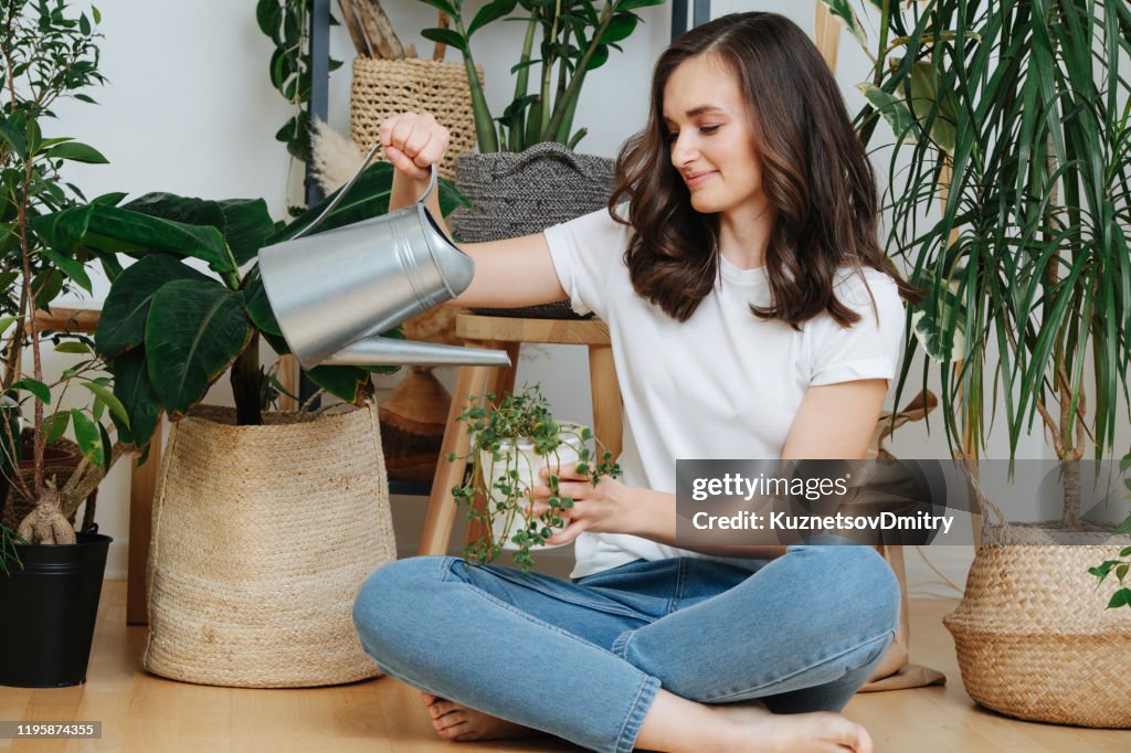 Young brunette sitting on a floor, watering potted plants in her home greenhouse