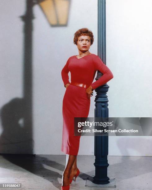 Eartha Kitt , US actress and singer, wearing a red dress, with a red belt, leaning against a lamppost in a studio portrait, the shadow of the...