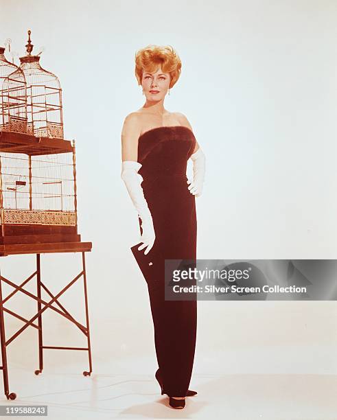 Eleanor Parker, US actress, wearing a long deep red strapless dress and long white gloves, while posing beside a birdcage in a studio portrait,...