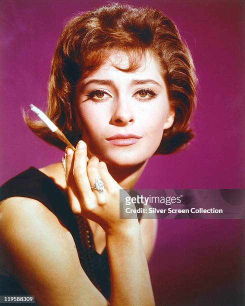 Elizabeth Ashley, US actress, poses with a lit cigarette, held in a holder, in a studio portrait, against a purple background, circa 1965.