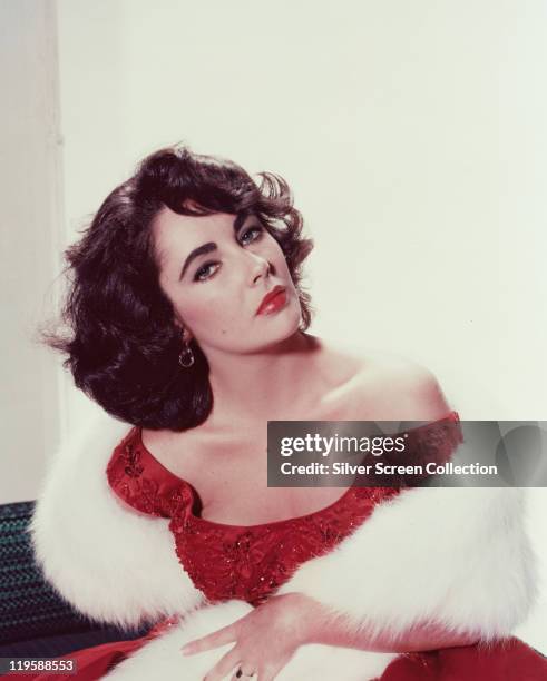 Elizabeth Taylor , British actress, wearing a red dress with a white fur stole in a studio portrait, against a white background, circa 1955.